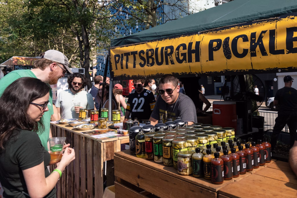 A person selling pickles at a market with several customers checking out the various products.